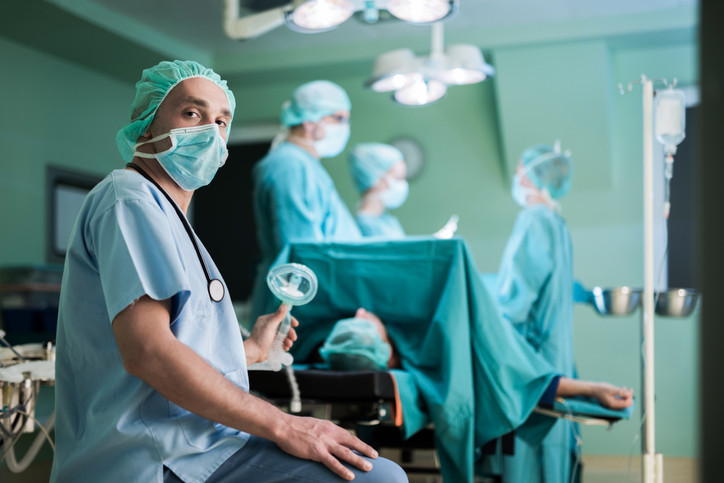 Male doctor sitting in operating room during surgery while holding oxygen mask and looking at the camera.