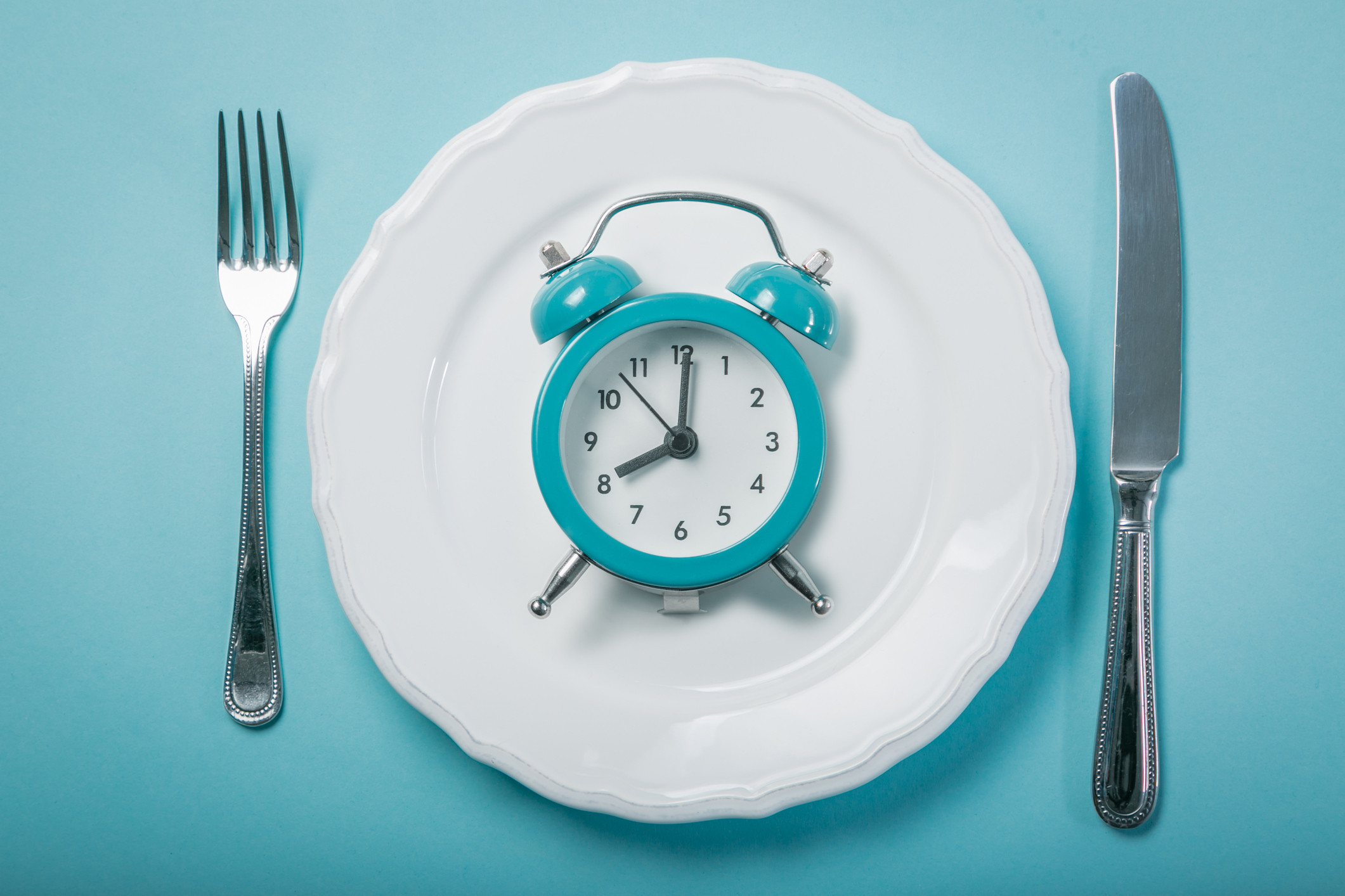 A plate surrounded by a knife and fork with a clock sitting on it. 