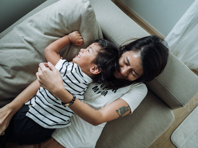 Smiling mother and young child lying down on a couch, mother has arm around child, who is laughing