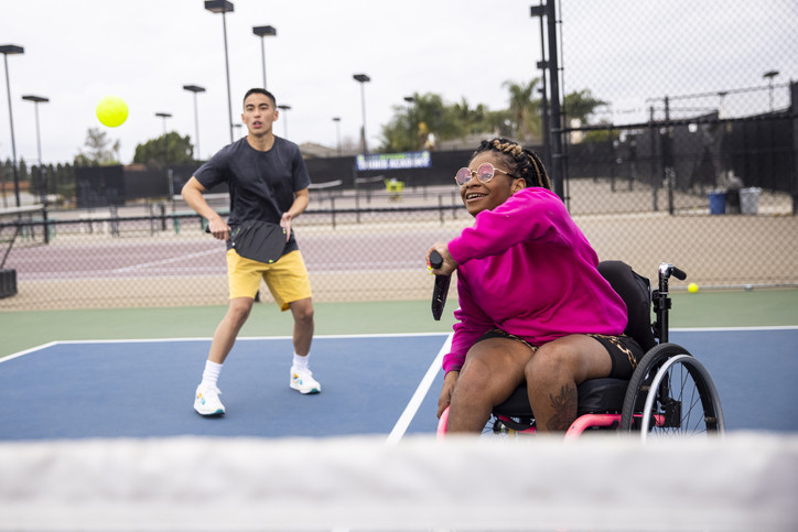 Un homme sur un terrain de pickleball vêtu d'un tee-shirt foncé et d'un short jaune et une femme en fauteuil roulant balançant sa raquette pour frapper la balle ;  elle porte un haut rose vif 