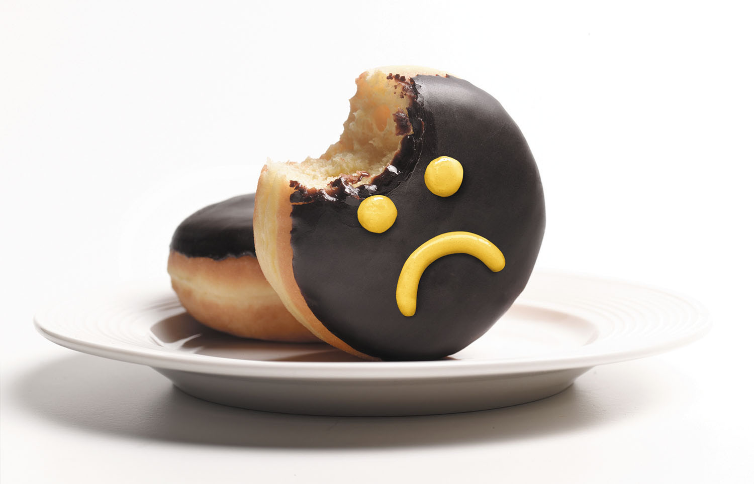 photo of two doughnuts on a plate, one is set on its edge, has a bite taken out of it, and has a sad face drawn on it in icing