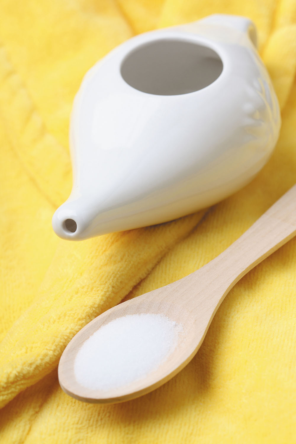 photo of a white neti pot with a wooden spoon next to it holding salt, both resting on a yellow cloth