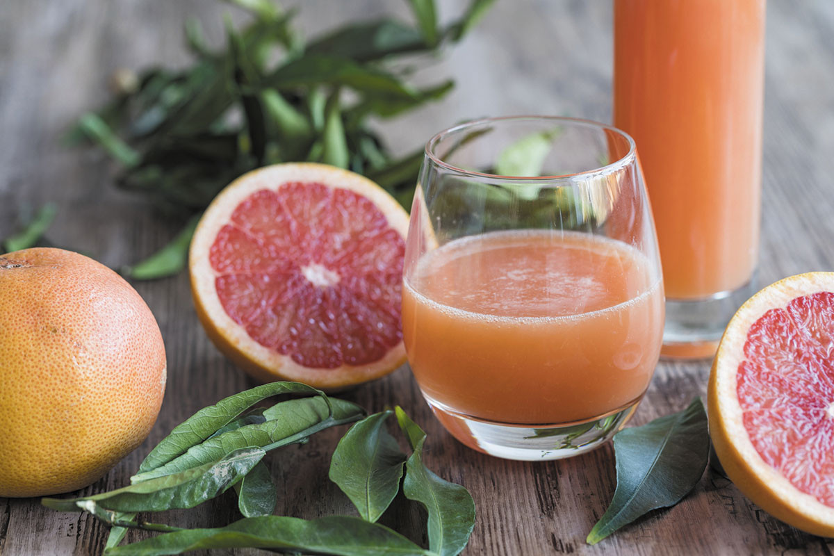 photo de pamplemousse coupé en deux sur une table à côté d'un verre à moitié plein de jus de pamplemousse, avec un pichet plein de jus derrière