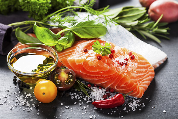 photo of a piece of salmon displayed with fresh herbs, a lemon, a chili pepper, and a dish of olive oil