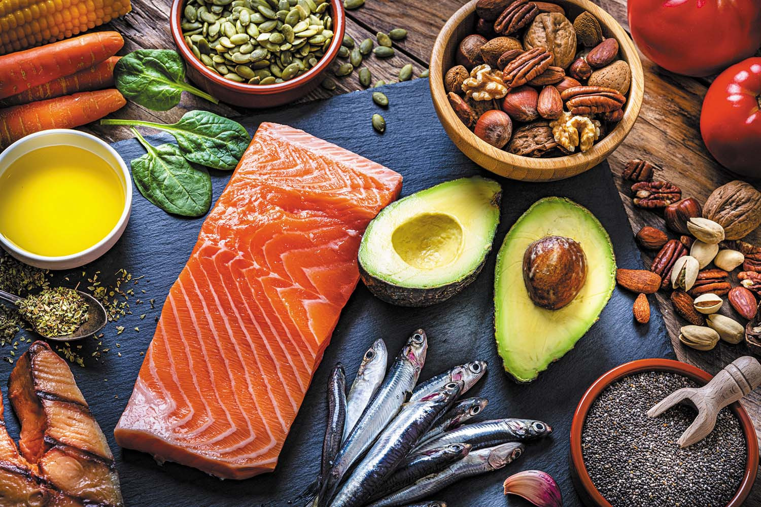 photo of an assortment of healthy foods: salmon, avocado, anchovies, spinach, nuts, olive oil, carrots, tomatoes