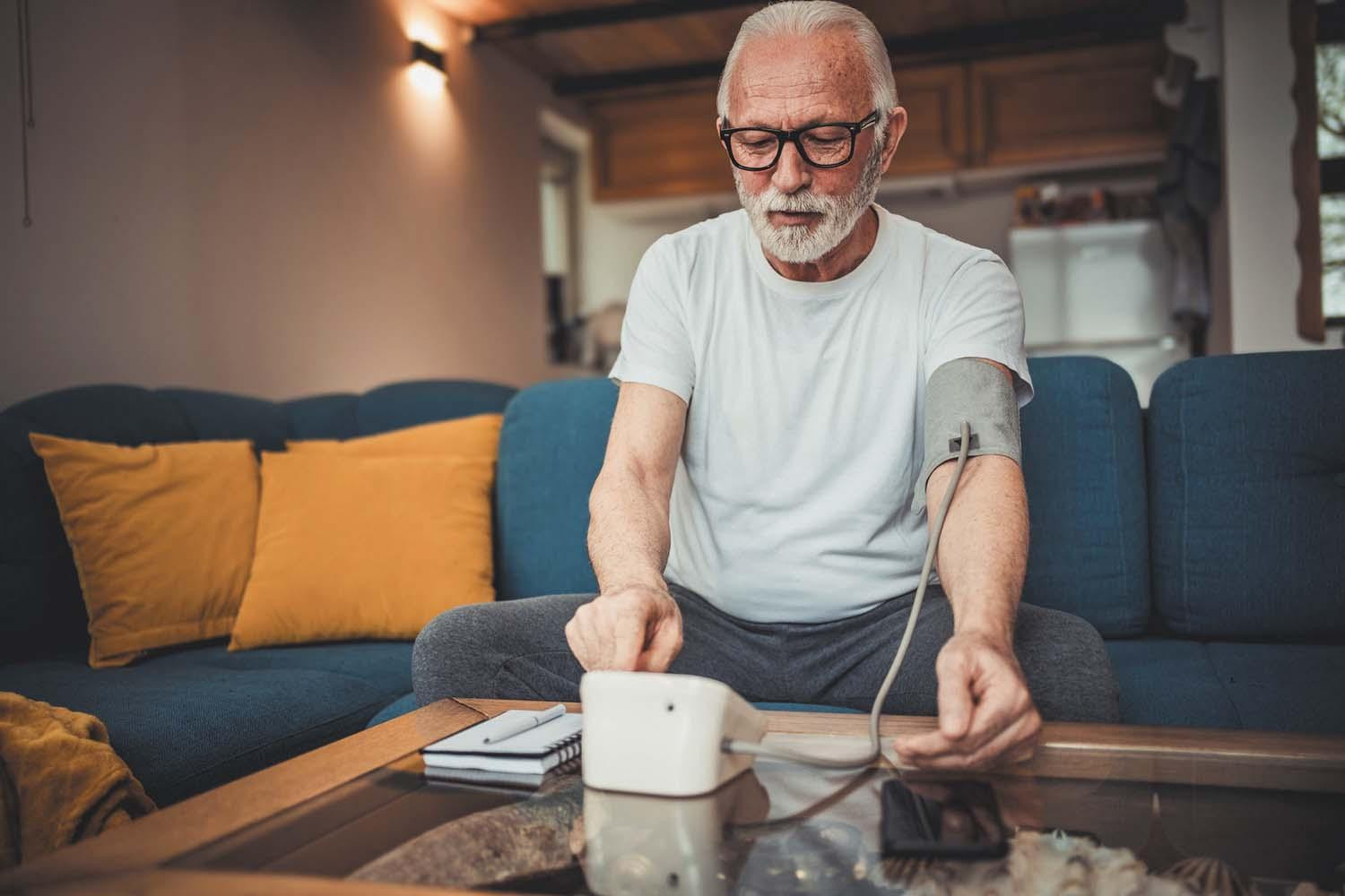 A senior man measures his blood pressure while sitting on the couch in his home.