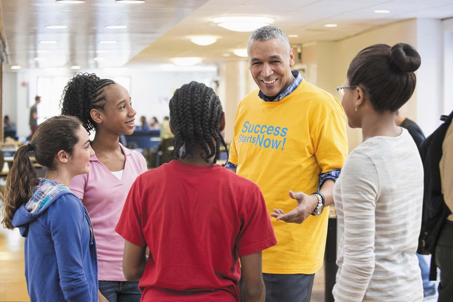 A senior man stands among a group of school-age children, talking to them as a mentor.