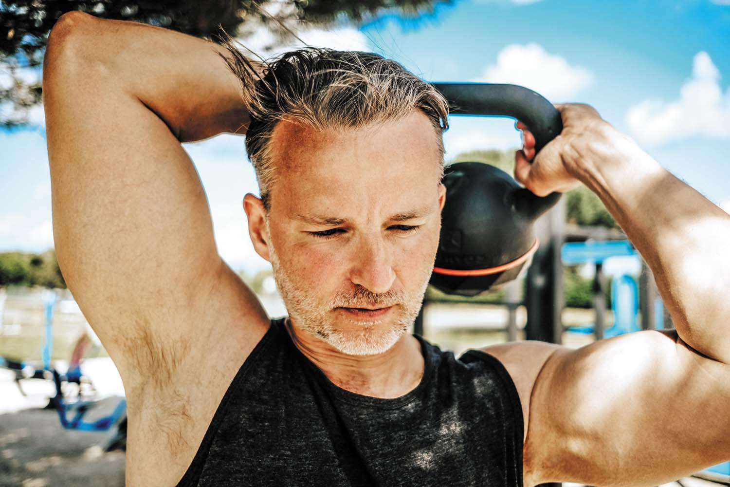 A mature man works out with a kettlebell, passing it behind his head.