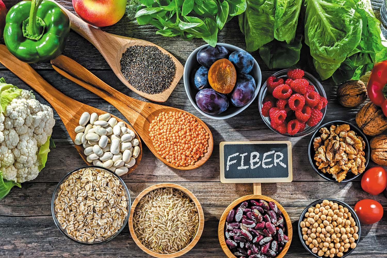 Overhead view of assorted high-fiber foods in bowls and serving spoons: berries, nuts, seeds, legumes, cauliflower, plums.