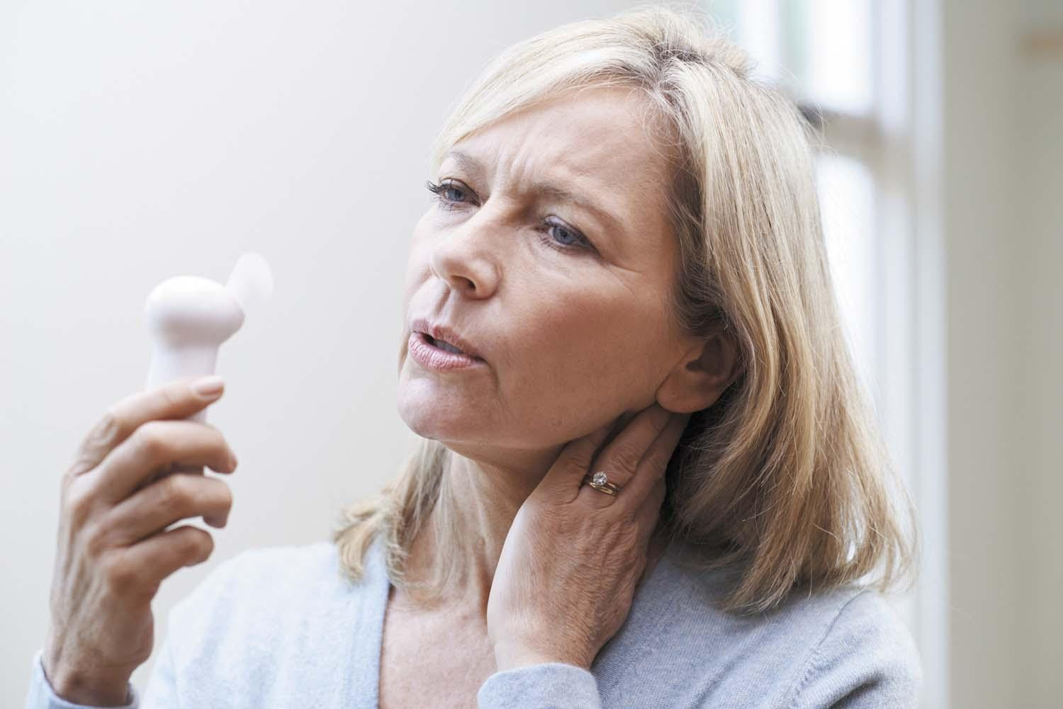 A mature woman holds a personal fan in front of her face, trying to cool down during a hot flash.
