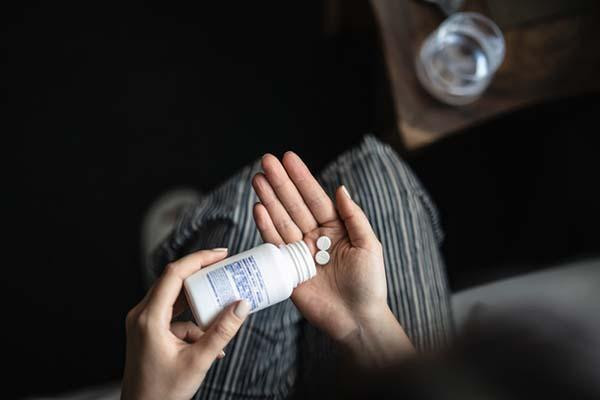 Overhead view of the hands of a person holding a medication bottle in one hand, tipping out two pills into the other hand; a glass of water is visible to the side, out of focus.