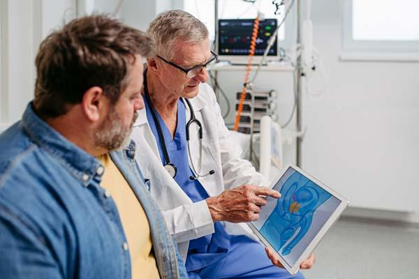 A doctor uses a tablet to show a middle-age male patient an illustration of male anatomy including the prostate gland, which is highlighted in yellow on the screen.