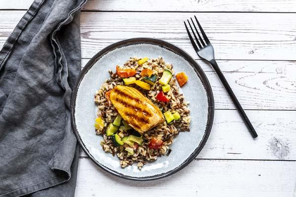 Overhead view of a dish of roasted salmon atop a mix of wild rice and diced vegetables; the plate is on a wood surface next to a fork and a cloth napkin.