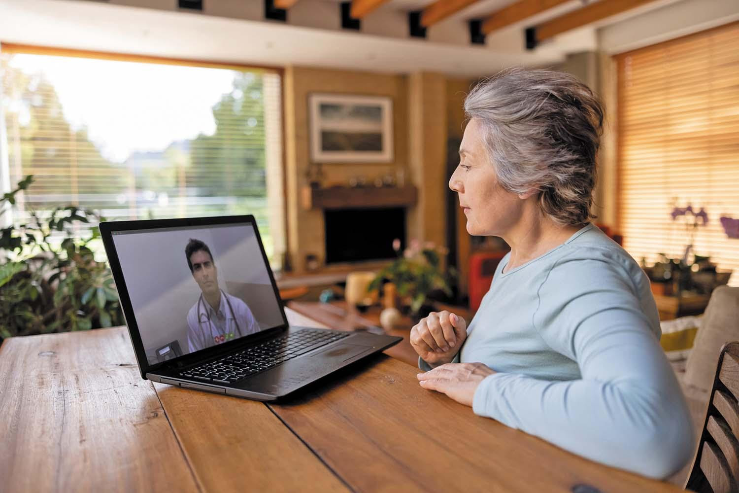 A woman sits in her home, using a laptop for a remote therapy session (a doctor is visible on the screen).