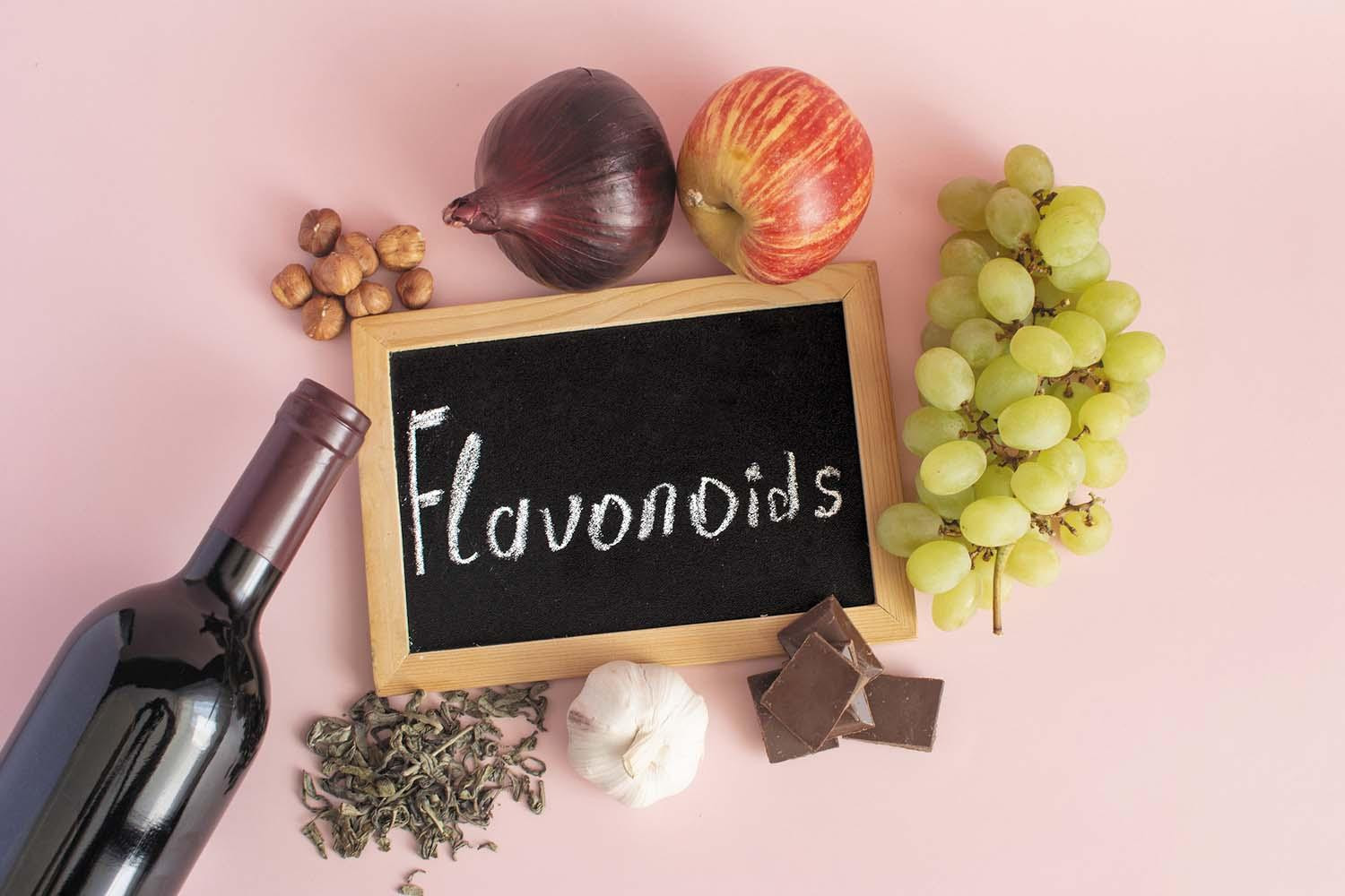 An assortment of foods rich in flavonoids (grapes, apple, onion, garlic, wine, dark chocolate) surrounding a small chalkboard with the word flavonoids written on it.