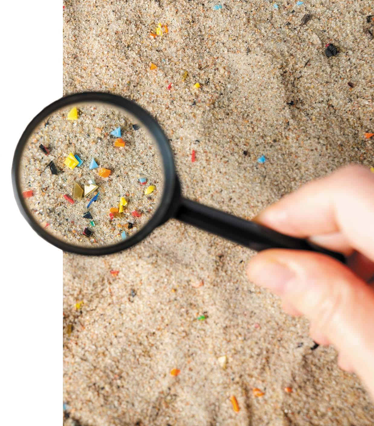 A magnifying glass held over sand shows particles of microplastics in assorted colors.