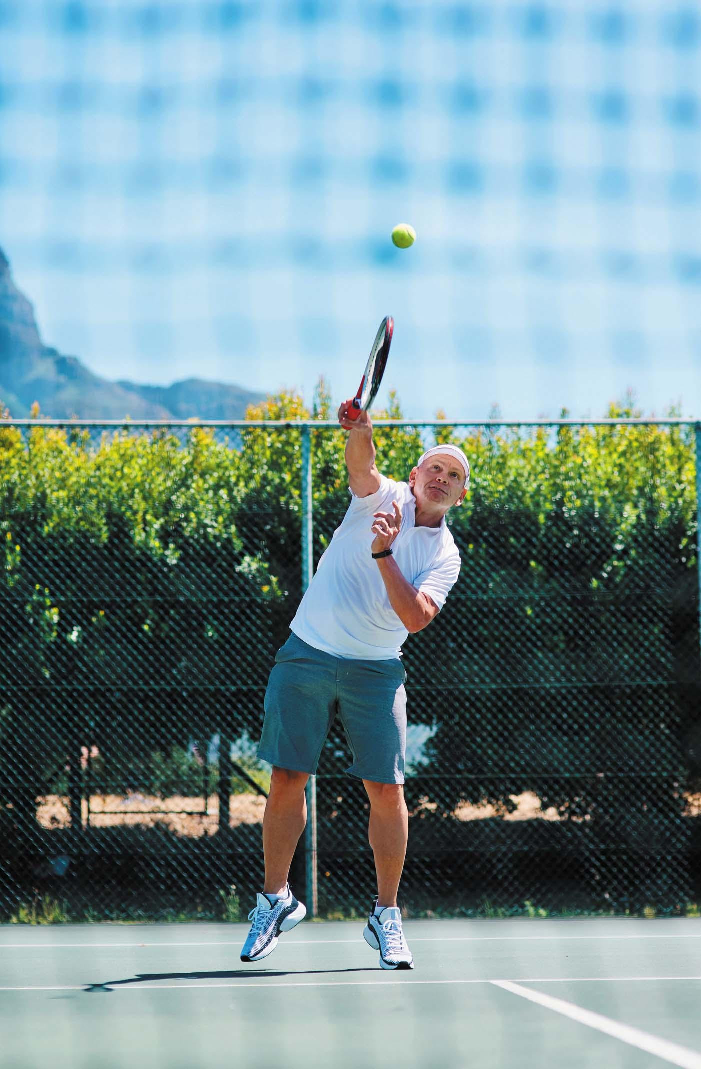 A senior man concentrates on a tennis ball just after he has hit it on an outdoor court.