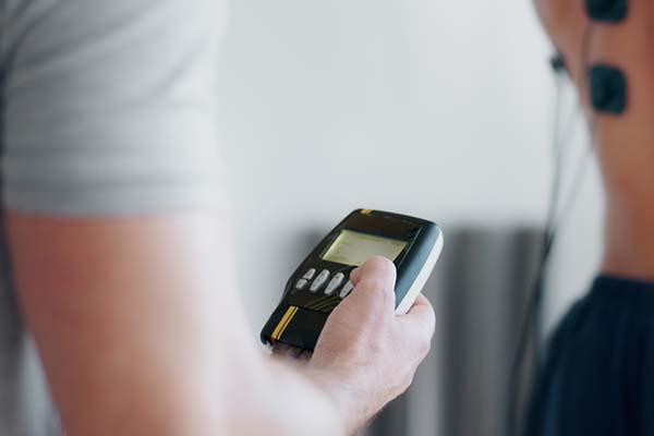 Cropped image of a man's torso in a white t-shirt; one hand is visible holding a biofeedback monitor.