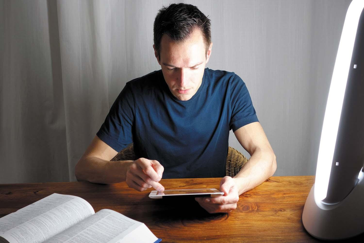 A man uses a tablet while sitting next to a light box used to treat seasonal affective disorder.