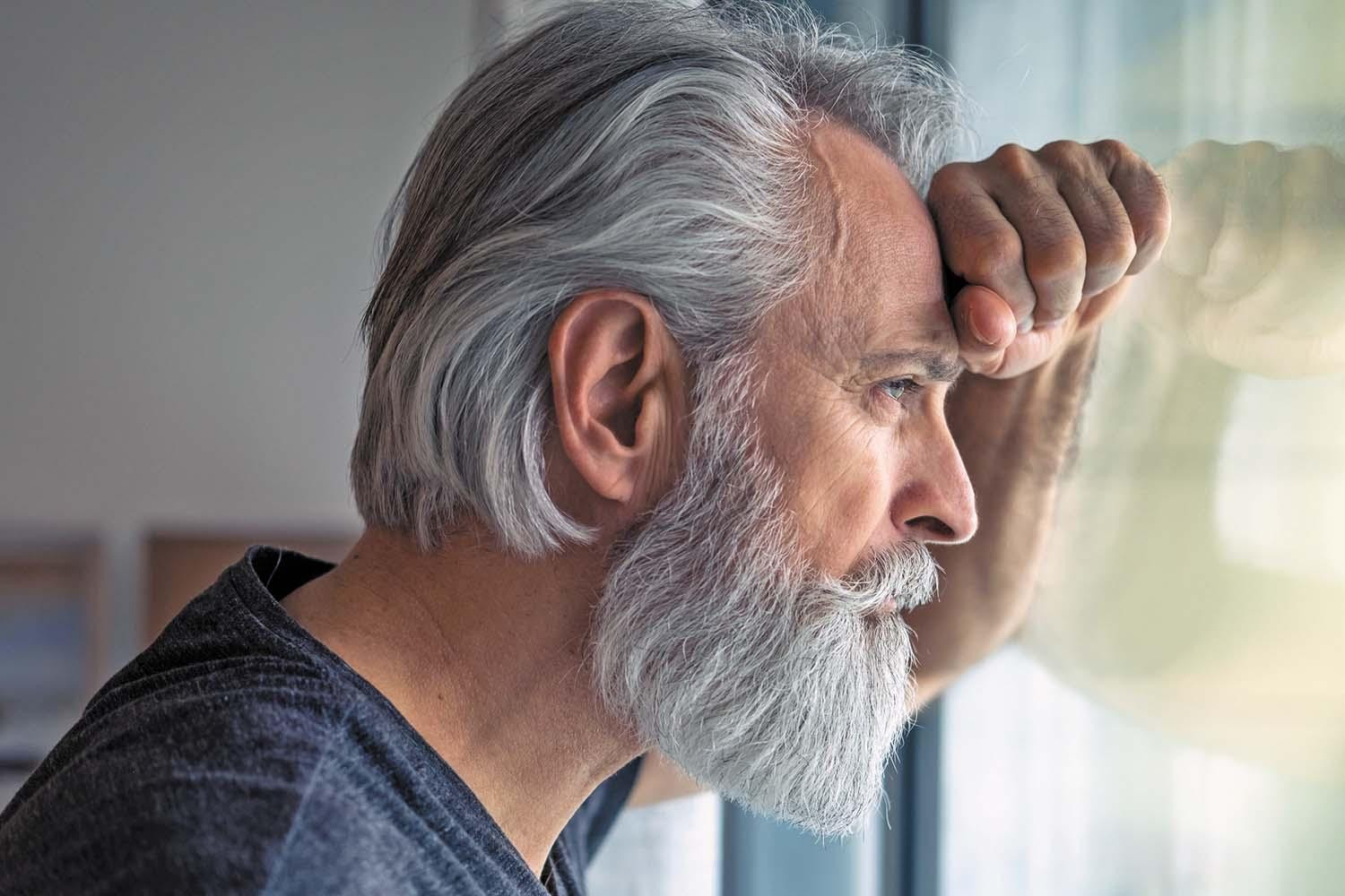 A mature man stands looking out a window with his forehead resting on his arm, which is against the glass.