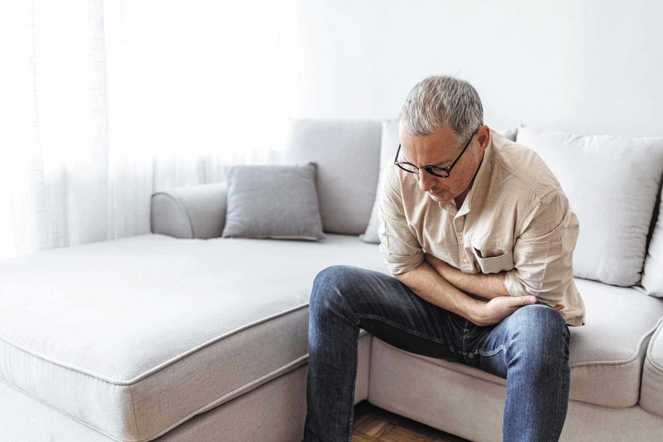 A man sits on a couch holding his arms against his lower abdomen as if in pain.
