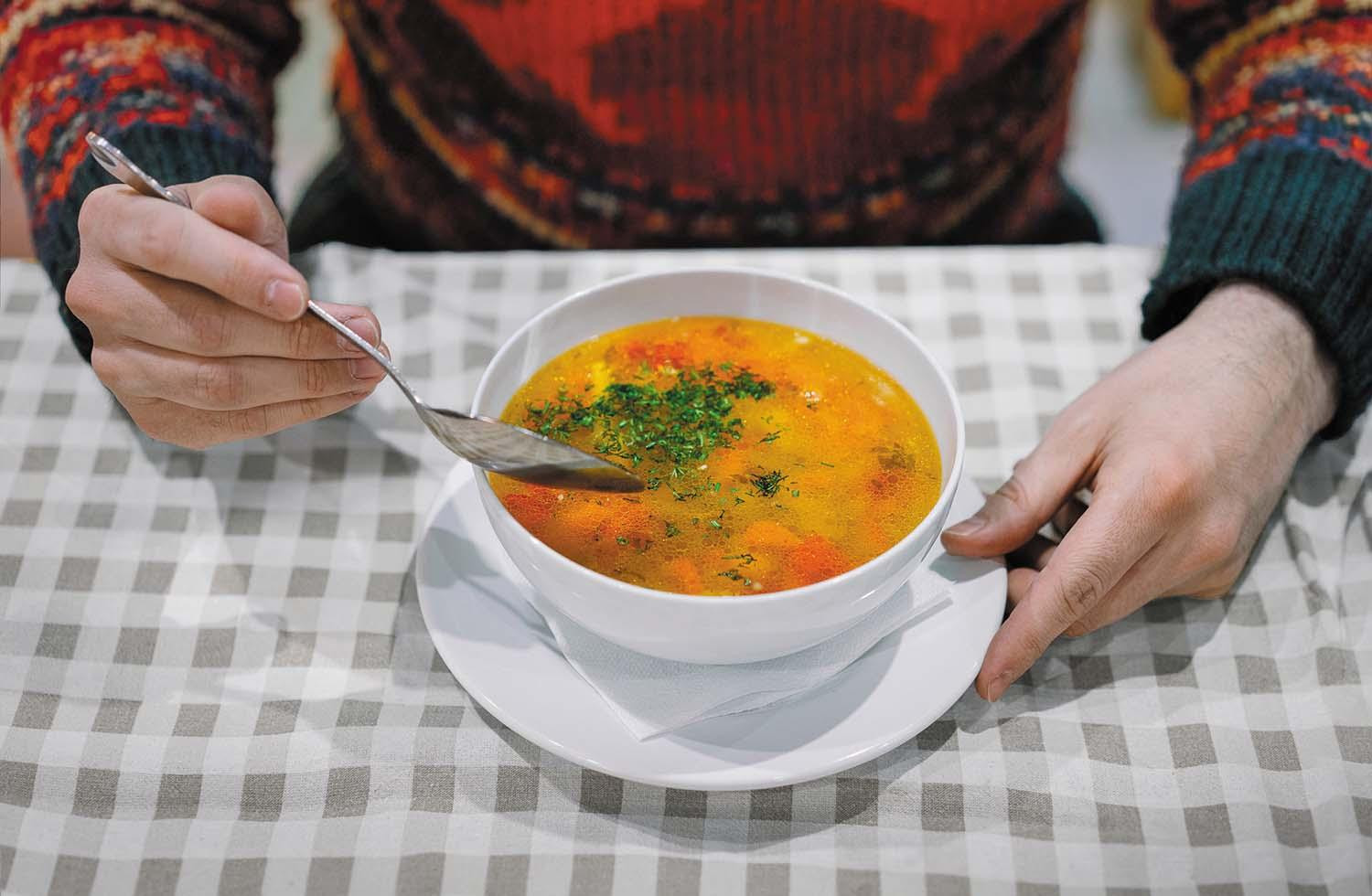 Cropped image shows torso and arms of a person sitting at a table eating a bowl of vegetable soup.