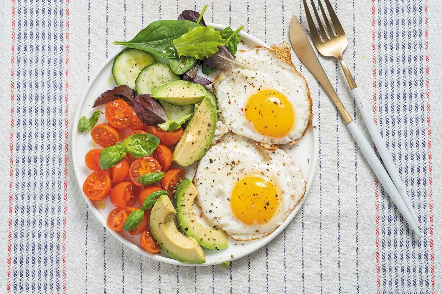 Breakfast of two fried eggs alongside a salad of lettuce, tomato, avocado, and cucumber.
