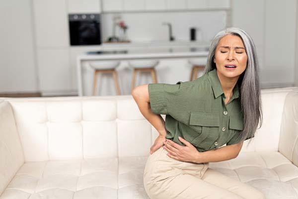 A mature woman sitting on a couch, holding a hand on her lower back and grimacing in pain.