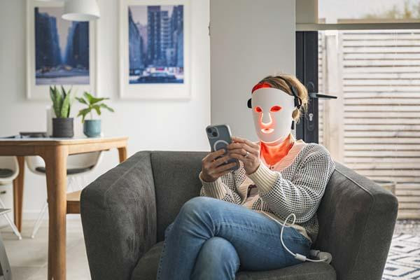 A woman uses her red LED face mask for red light therapy while relaxing at home in and using her smartphone.
