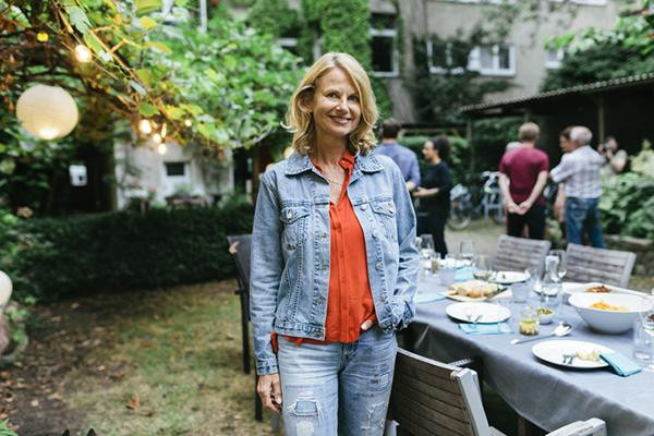 A middle-age woman smiles as she stands next to a table in a back yard; several people are gathered behind her, out of focus.