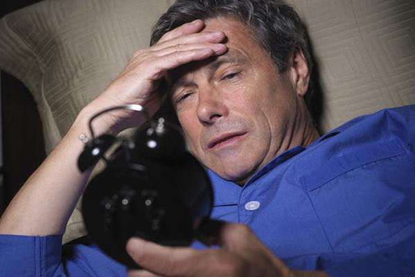 A man in bed wearing pajamas looks at an old-fashioned alarm clock with bells on top.