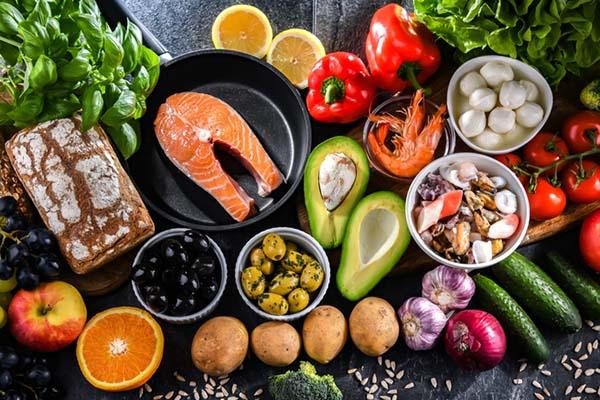 An assortment of food products representing the Mediterranean diet: salmon, shrimp, avocado, garlic, peppers, olives, whole-grain bread, citrus fruits, apples, grapes, tomatoes, and more.