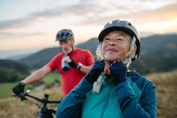 A mature couple on a bike ride taking a break, the woman fastens helmet in the foreground as the man drinks water behind her, out of focus.