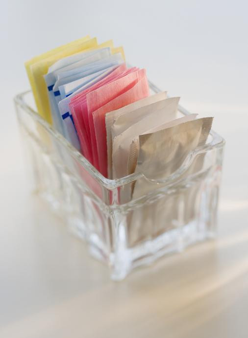 A small glass dish holds packets of artificial sweeteners in different colors: yellow, blue, pink, white.
