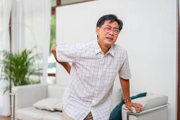 A middle-age man experiencine back pain, leaning on a chair with one hand and holding the other to his lower back.