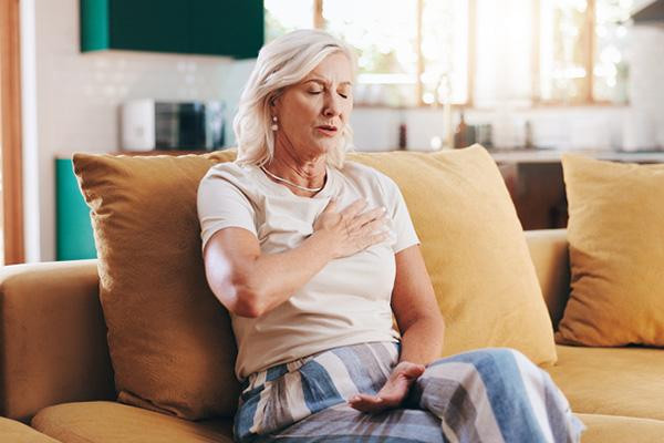 A senior woman sitting on a couch in her home holds her hand to her chest with her eyes closed, experiencing chest pain.