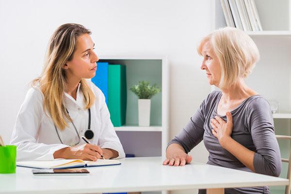 A patient and a doctor sit facing each other at a table; the patient has a hand on her chest, describing chest pain.