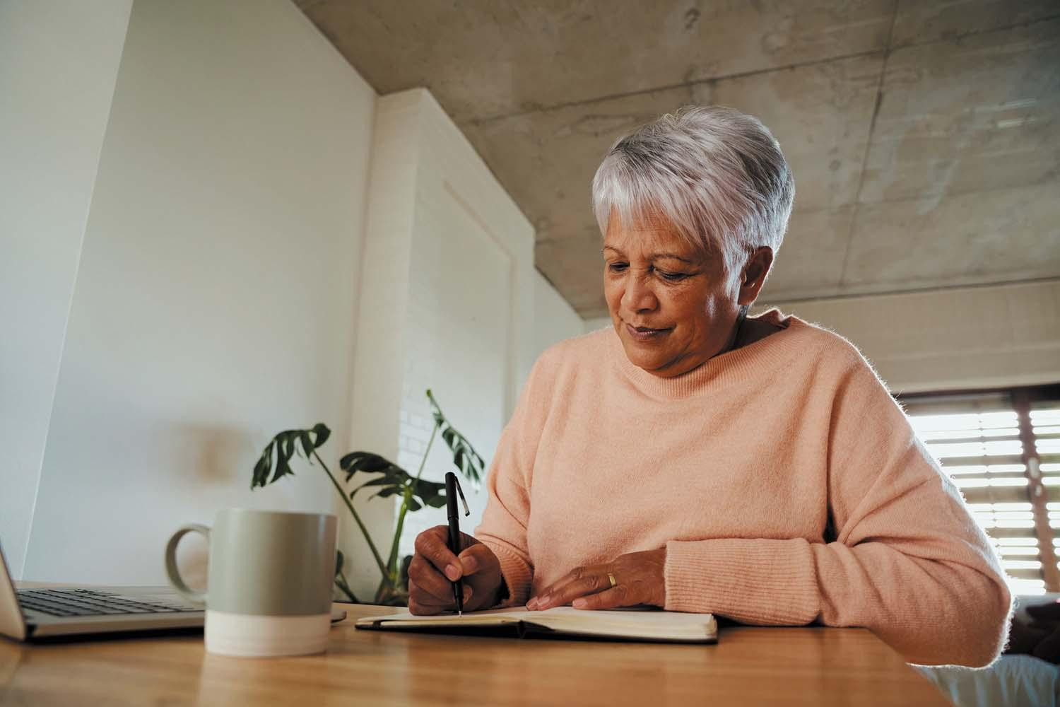 A senior woman sits at a table, writing information in an appointment book.