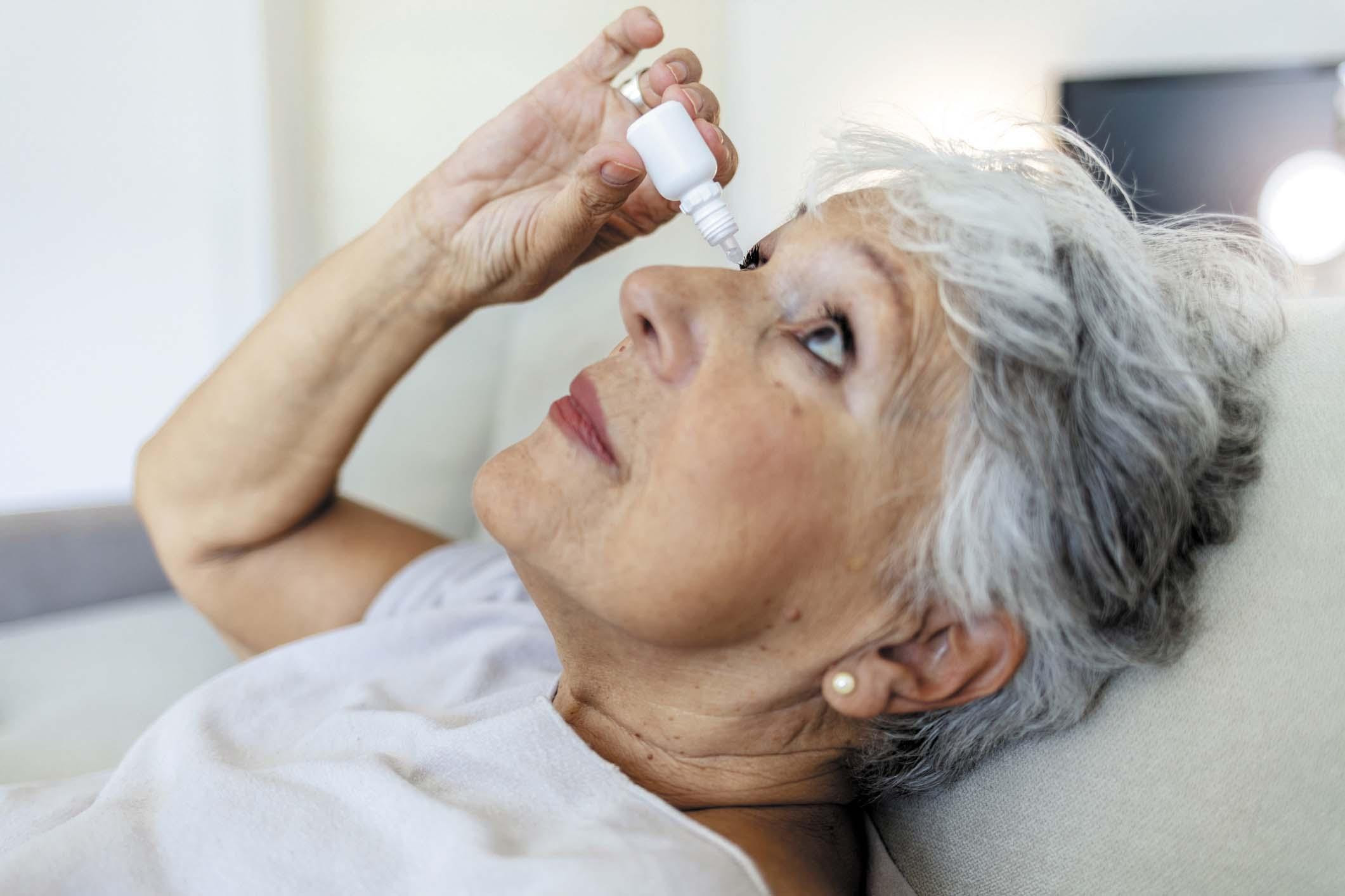 A mature woman leans back while holding a bottle of eye drops above one eye.