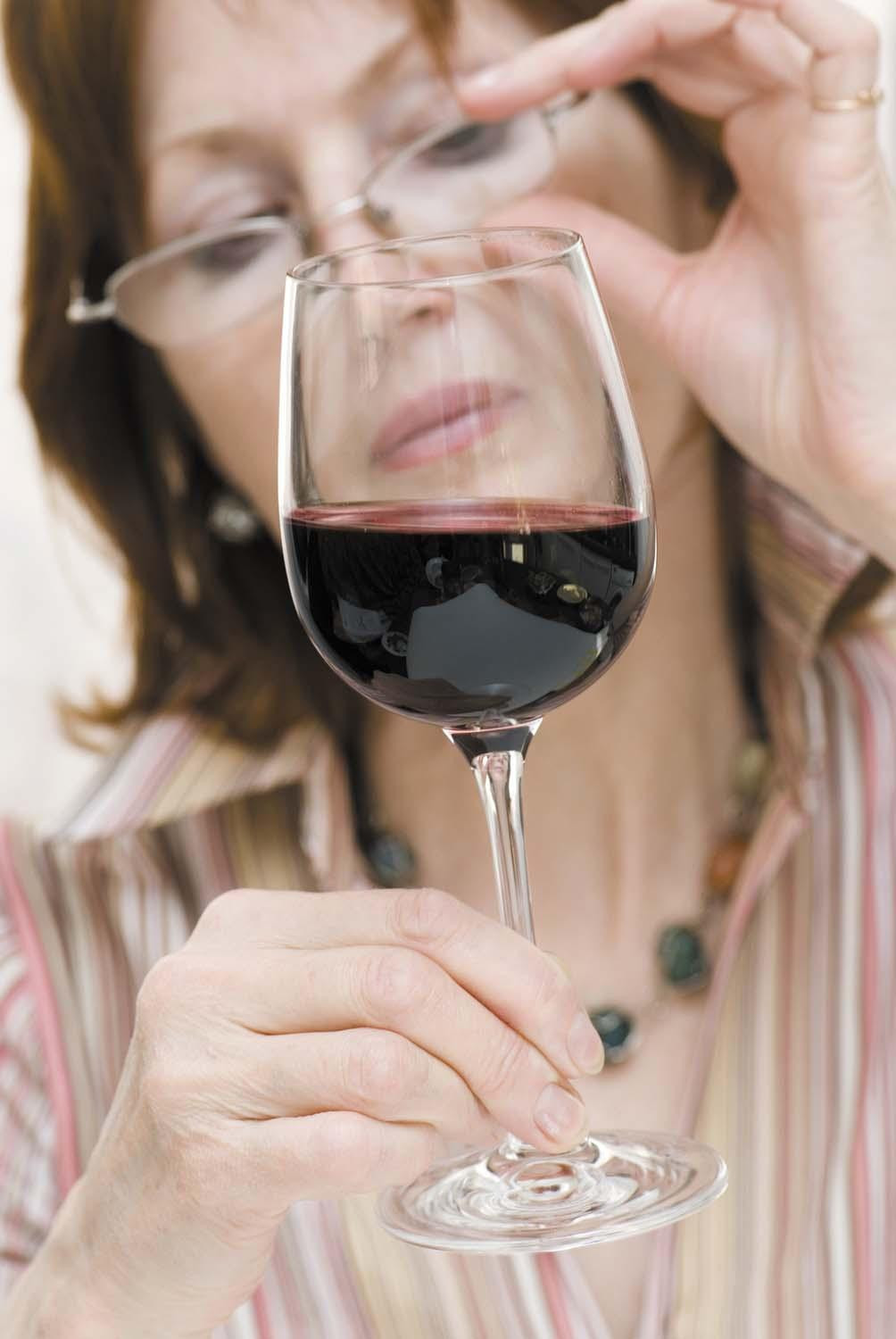 A woman holds a glass of red wine in front of her face while adjusting her glasses.