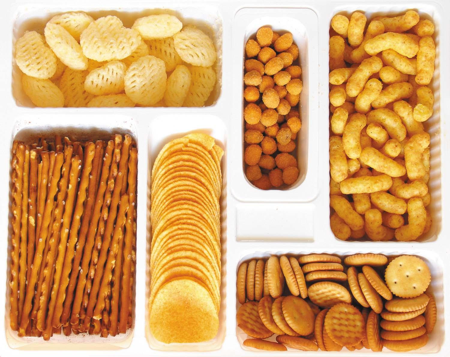 A white tray holds various kinds of ultraprocessed foods in individual sections: crackers, cheese puffs, stacking potato chips, pretzels, and others.