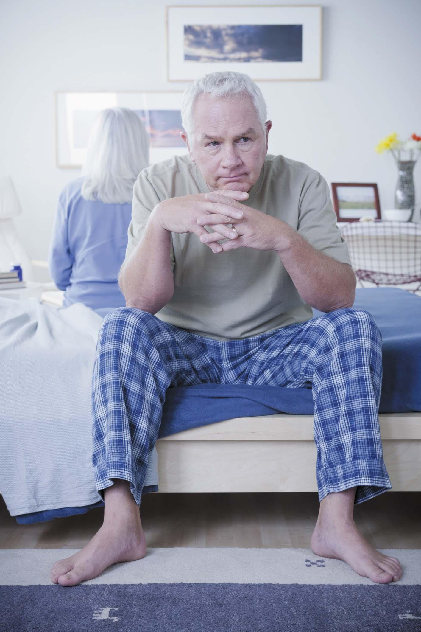 A mature couple sitting on opposite sides of a bed; the man is facing the camera and looks upset and disappointed.