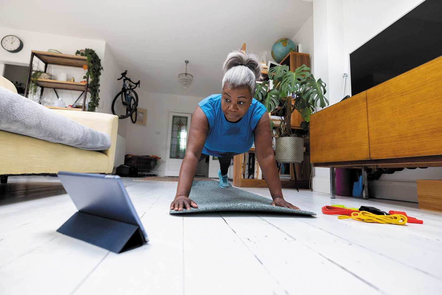 A mature woman in position to do a push-up while viewing a tablet; she is on an exercise mat in her home.