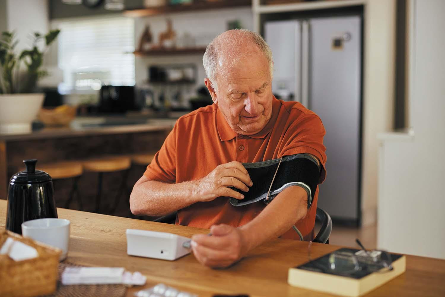 A senior man sitting in a kitchen attaches an arm cuff so he can measure his blood pressure at home.