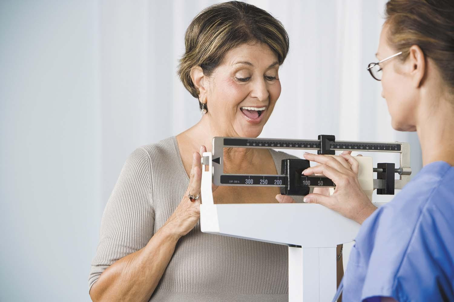 A happy mature woman has her weight measured in a doctor's office.