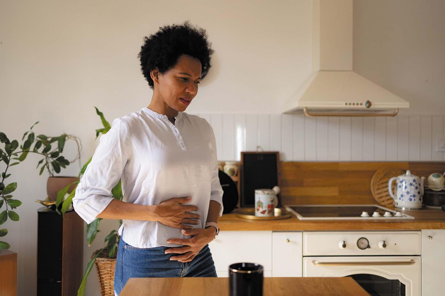 A woman stands in her kitchen, holding both hands to her stomach indicating discomfort.