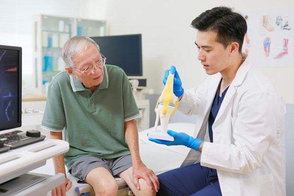 A doctor uses a model of a human knee to explain joint replacement to a senior man in an exam room.