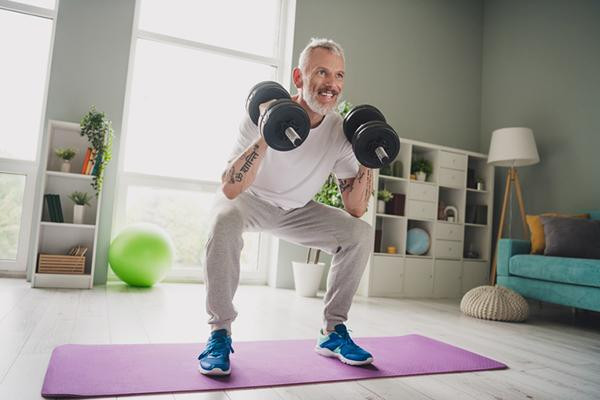 A mature man exercises with dumbbells in a space in his home, with light coming through large windows behind him.
