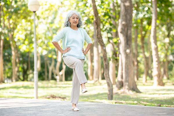 A senior woman stands outdoors with her hands on her hips, raising one leg to test her balance.