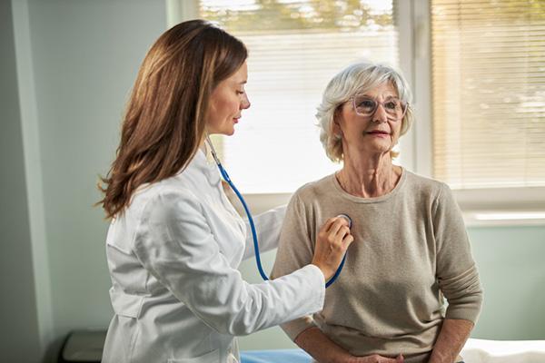 A doctor uses a stethoscope to listen to the heartbeat of a seated senior woman.